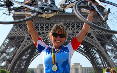 Female cyclist holing a bike above her head, with a medal around her neck, in front of the Eifel Tower