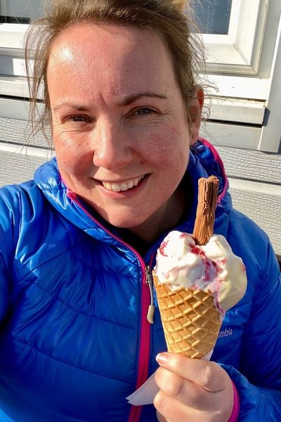 Clare eating a soft-scoop ice cream in a cone with flake, Clare is wearing a blue coat and smiling at the camera.