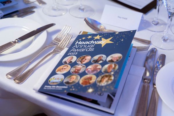 Headway Annual Awards 2023 brochure showing finalists faces in circles on blue background. Brochure is placed on a crisp white table cloth with formal place setting.