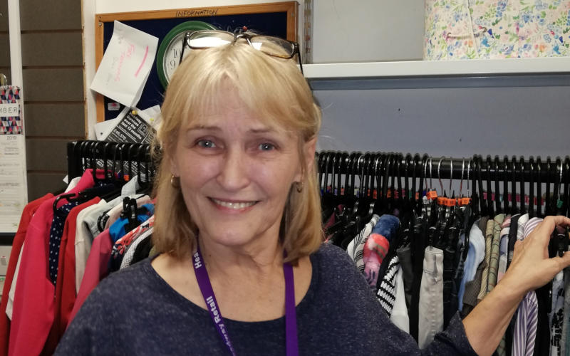 Jayne Copeman, Headway charity shop volunteer, stands smiling in front of a rail of clothes.