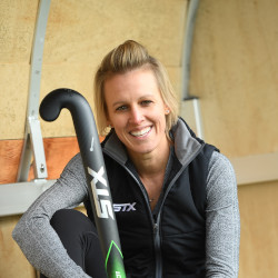 Alex Danson sits smiling at the camera holding a hockey stick.