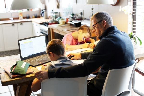 Two boys being home schooled by their Dad. The children are writing on notepads sitting at a table in their kitchen. The Dad is supervising one of the children, who also has work open on a laptop that is placed in front of him.