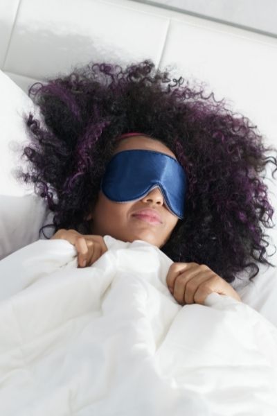 Woman sleeping in bed, it's daylight in the room and she is wearing a blue eye mask.